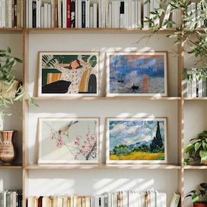 May include: Four framed art prints displayed on a wooden bookshelf. The prints feature various scenes, including a woman relaxing, a sunset over water, a bird on a flowering branch, and a landscape with a cypress tree.