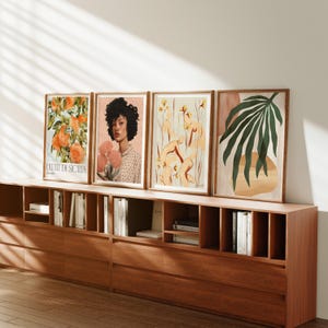 May include: Four framed art prints on a wooden shelf. The prints feature a woman with dark hair, a still life of fruit, a floral design, and a palm leaf.