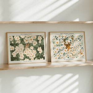Könnte beinhalten: Zwei gerahmte Drucke mit weißen Blumen und grünem Laub. Der Druck auf der linken Seite zeigt ein kleines Eichhörnchen, das aus den Blumen hervorlugt. Der Druck auf der rechten Seite zeigt ein Reh mit Geweih, das in den Blumen versteckt ist. Beide Drucke haben den Text "The Secret Garden" oben und "You've got a secret of gold" unten.