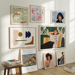 Könnte beinhalten: Eine Galeriewand mit gerahmten Kunstdrucken in verschiedenen Stilen. Die Sammlung umfasst florale, abstrakte und Porträtkunst. Die Rahmen sind aus hellem Holz, und die Kunst wird an einer weißen Wand über einem Holzboden ausgestellt. Ein kleiner Holzschemel mit Büchern befindet sich im Vordergrund.