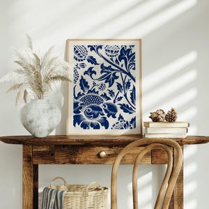 May include: A framed print of a blue and white floral pattern. The print is on a white background and features a repeating pattern of stylised flowers and leaves. The print is hanging on a wall above a wooden table.