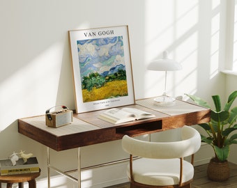 May include: A framed print of a painting by Vincent van Gogh titled "Wheat Field with Crows" depicting a yellow wheat field with a blue sky and crows flying overhead. The print is on a wooden desk with a book, a lamp, and a small radio.