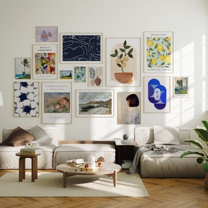 May include: A living room interior with a white wall featuring a gallery wall of framed art prints. The prints include abstract art, landscapes, and still life. The room is furnished with a white sectional sofa, a coffee table, and a plant.