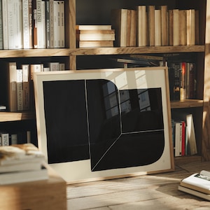 Peut inclure: Une impression d'art encadrée avec un design géométrique noir audacieux sur un fond crème. L'œuvre est présentée sur un sol en bois devant une bibliothèque remplie de livres. Le cadre est de couleur bois clair.