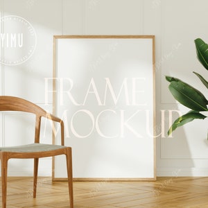 May include: A wooden chair with a light gray cushion sits in front of a white wall with a large, empty picture frame. A potted green plant is on the right side of the image.