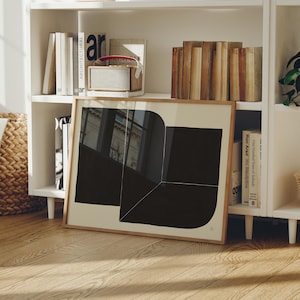 May include: A framed abstract art print with a black and white geometric design. The artwork is displayed on a wooden floor in front of a white bookshelf filled with books and decorative items. The print has a light brown wooden frame.