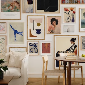 May include: A gallery wall featuring framed art prints in various styles and sizes. The artwork includes abstract designs, portraits, and floral arrangements. The room also features a white sofa, wooden table, and chairs, creating a stylish interior.