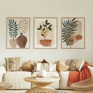 Könnte beinhalten: Drei minimalistische abstrakte Kunstdrucke mit grünen Blättern, einer Topfpflanze und geometrischen Formen in Braun-, Beige- und Gelbtönen. Die Drucke sind an einer weißen Wand über einem beigen Sofa mit braunen und beigen Kissen und einem geflochtenen Couchtisch angebracht.
