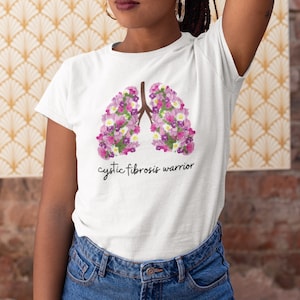 May include: White t-shirt featuring a floral lung design in shades of pink, purple, and white, with brown stems. The text "cystic fibrosis warrior" is printed below the graphic. The shirt is paired with blue jeans.