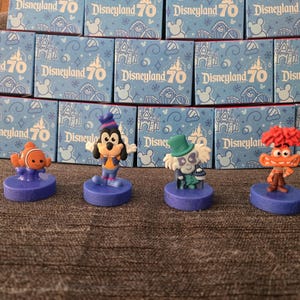 Puede incluir: Cuatro figuras de personajes sobre bases moradas se exhiben frente a cajas que dicen "Disneyland 70". Las figuras incluyen a Nemo, Goofy, un fantasma y un personaje con cabello naranja. Las cajas son de color azul claro con un diseño de castillo.