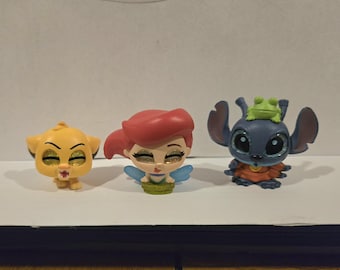 Könnte beinhalten: Drei Disney-Charakterfiguren: Simba, Arielle und Stitch. Simba ist gelb, Arielle hat rotes Haar und blaue Flügel, und Stitch ist blau mit einem Frosch auf dem Kopf. Alle haben glitzernde Augen und stehen auf einer weißen Oberfläche.