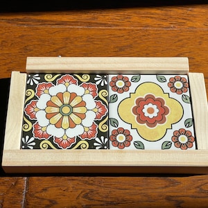May include: A decorative tile coaster with two square tiles featuring floral designs in orange, yellow, and red. The tiles are set in a light wood frame, creating a vibrant and functional home decor item.
