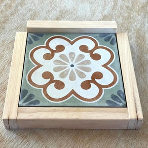 May include: Square wooden trivet with a decorative tile insert. The tile features a floral design in white, brown, and green, set within a light wood frame. The trivet is designed to protect surfaces from heat.