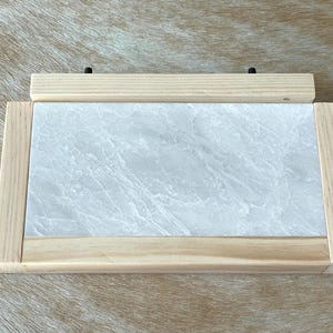 May include: A rectangular wooden picture frame with a light-colored marble-like insert. The frame is made of light-colored wood and has a simple, clean design. Two black screws are visible on the top wooden piece.