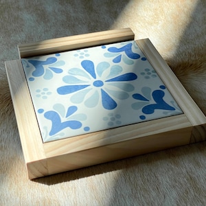 May include: Square wooden tray with a floral tile insert. The tile features a light blue and white floral pattern. The wooden frame is made of light-colored wood. The tray is sitting on a textured surface.