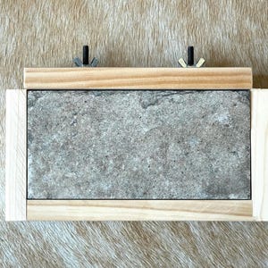 May include: A rectangular gray stone tile is framed by light-colored wood. Two bolts with wing nuts secure a wooden bar to the top. The item is likely a trivet or a similar kitchen or home decor item.