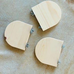 May include: Three unfinished, semi-circular wooden shelves with attached metal hardware. The shelves are light-colored with visible wood grain. Each shelf has two metal wing nuts for attachment. The background is a light brown, textured surface.