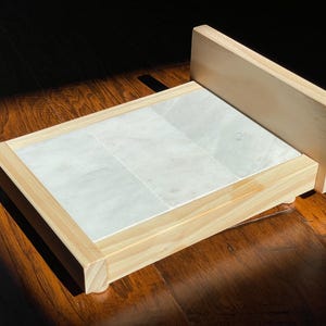 May include: A wooden serving tray with a light-colored marble tile insert. The tray has a raised back and is made of light-colored wood. The marble tiles are rectangular and arranged in a grid pattern. The tray is sitting on a dark wood surface.