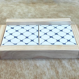 May include: Wooden serving tray with two white tiles featuring a blue lattice pattern. The tray has a raised back and is made of light-colored wood, suitable for serving appetizers or small dishes.