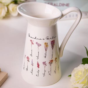May include: White ceramic jug with a handle, featuring a floral design and the text "Grandma's Garden". The jug has a variety of flowers and names written on it.