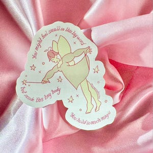 May include: A sticker featuring a whimsical fairy illustration in soft pastel colors. The fairy is depicted with a wand and wings, surrounded by stars. The sticker has text that reads, "You might feel small in this big world but inside this tiny body you hold so much magic."