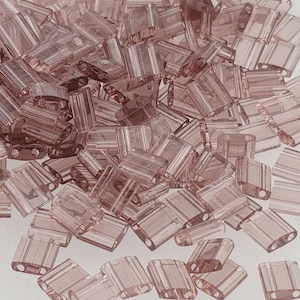 Könnte beinhalten: Ein Haufen transluzenter, rechteckiger Perlen in einem sanften Malvenfarbton. Jede Perle hat eine rechteckige Form mit abgerundeten Kanten und zwei Löchern zum Auffädeln. Die Perlen sind verstreut und bilden eine strukturierte Oberfläche.