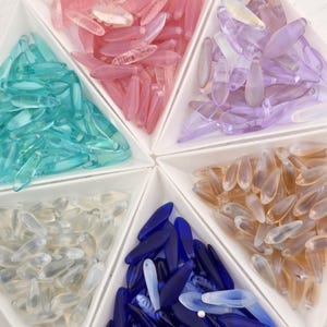 May include: A collection of teardrop-shaped beads in various colors, including pink, teal, purple, clear, amber, and blue, are arranged in a white segmented tray. The beads are translucent and appear to be made of glass or acrylic.