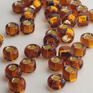 Puede incluir: Un conjunto de cuentas de vidrio color ámbar con interior dorado. Las cuentas son cilíndricas con bordes redondeados y un agujero en el centro. Las cuentas están dispersas sobre una superficie blanca.