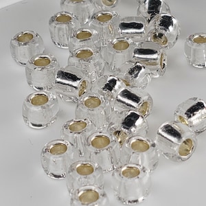 May include: A collection of small, barrel-shaped beads featuring a clear, transparent glass exterior and a reflective, metallic silver-colored core. Each bead has a central hole for threading, with a smooth, polished finish throughout the set.