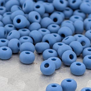 May include: A collection of small, round, matte-finish beads in a solid dusty blue color. Each bead features a central hole for threading. The beads are scattered across the surface, showing a uniform texture and consistent light blue hue throughout.
