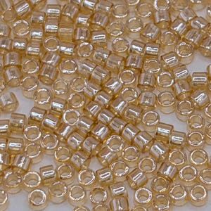May include: A close-up shot of a pile of small, cylindrical beads. The beads are a translucent, light amber color, with a slightly iridescent sheen. They are evenly shaped and appear to be made of glass or a similar material, suitable for jewelry making.