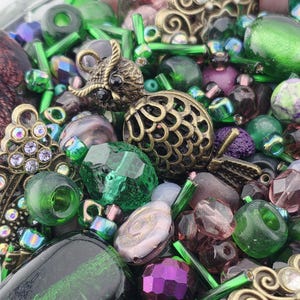 May include: A mix of beads and decorative elements in shades of green, purple, and gold. The assortment includes faceted glass beads, cylindrical beads, and ornate metal charms. The color palette is rich and varied, with a mix of opaque and translucent elements.
