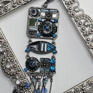 May include: A decorative keychain featuring a rectangular silver-toned metal frame with clear rhinestones. Attached is a multi-layered pendant composed of geometric black and silver-toned metal components, accented with bright blue rhinestones and beads.