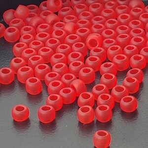 May include: A collection of numerous small, round, translucent red beads. Each bead features a smooth, matte finish and a central hole for threading. The beads are scattered across the surface, displaying a consistent vibrant red color throughout.