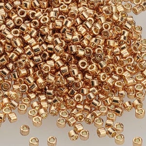 May include: A close-up shot of a pile of small, shiny, gold-colored beads. The beads are cylindrical with a hole running through the center, and they are scattered across a light-colored surface. These beads are ideal for jewelry making and crafting.