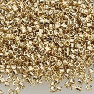 May include: Close-up of a pile of small, cylindrical gold-colored beads. The beads have a metallic sheen and are densely packed together, suggesting they are for jewelry making or crafts.