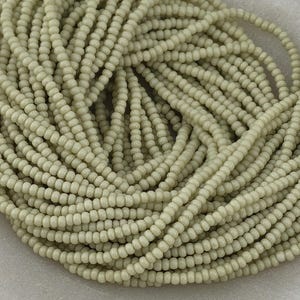 May include: A large collection of small, round, matte-finish beads in a soft, muted sage green color. The beads are strung together on multiple threads, creating a dense, coiled arrangement that fills the frame with a uniform, textured appearance.
