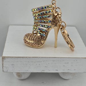 May include: Gold-colored high-heeled shoe keychain. The shoe is adorned with iridescent rhinestones and smaller brown rhinestones on the toe. The keychain has a gold ring and clasp.