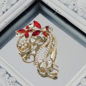 May include: A gold-toned brooch with red and clear crystals. The brooch features a floral design with red leaf-shaped crystals and clear, round crystals. The brooch is set against a reflective surface within a white, ornate frame.