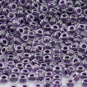 May include: A close-up shot of a pile of translucent purple glass beads. The beads are round with a hole through the center, and they reflect light, creating highlights. These beads are suitable for jewelry making and crafts.
