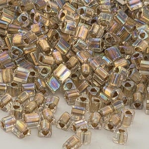 May include: A close-up shot of a pile of small, iridescent, tube-shaped beads. The beads have a translucent, golden-brown color with hints of rainbow iridescence. Each bead has a triangular hole at each end, suggesting they are for stringing.