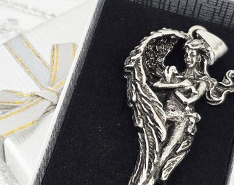 May include: A silver pendant in the shape of a winged figure, presented in a gift box. The pendant features intricate details and is set against a black background. A silver gift box with a silver and gold ribbon is in the background.