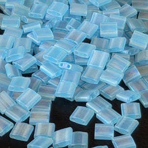 May include: A collection of light blue, rectangular glass beads with a translucent, iridescent finish. Each bead features two parallel holes running through the center, designed for stringing. The beads are scattered in a loose pile.