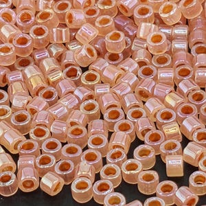 May include: Close-up of a pile of small, cylindrical beads in a soft peach color. The beads have a slightly iridescent finish, catching the light and creating highlights. The beads are uniform in shape and size, perfect for jewelry making and crafts.