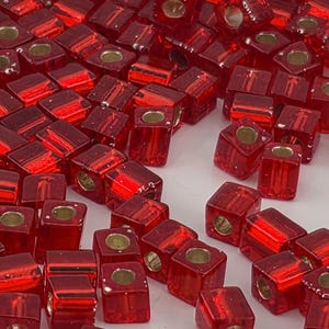 May include: A collection of small, translucent red glass beads, each shaped as a cube with a central circular hole. The beads feature a smooth, reflective surface and a consistent deep red color, scattered across the frame in a dense arrangement.
