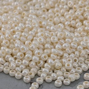 May include: A collection of small, off-white, pearlescent beads. The beads are round with a hole through the centre, and they have a shiny, reflective surface. The beads are scattered across a grey surface, creating a textured appearance.