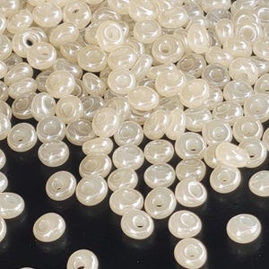 May include: A close-up shot of a pile of ivory-colored, round beads. Each bead has a central hole and a slightly iridescent sheen, suggesting a pearl-like appearance. The beads are scattered, creating a textured surface.