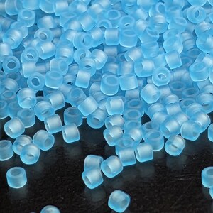 May include: A close-up shot of a pile of translucent, light blue, cylindrical beads. The beads are scattered across a dark surface, with a soft, frosted appearance. The beads are uniform in shape and size, ideal for jewelry making.