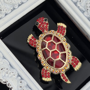 May include: A gold-toned turtle brooch with a red, faceted body and red and yellow gemstone accents. The brooch has black eyes and is set against a black background, framed by a white, ornate border.