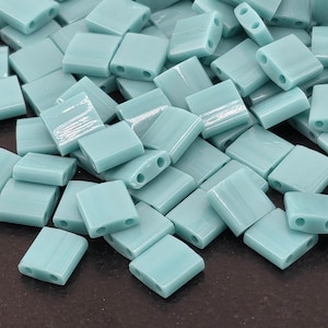 May include: A pile of light blue, rectangular beads with two holes each. The beads are a soft, opaque color and appear to be made of glass or a similar material. These beads are suitable for jewelry making and crafting.